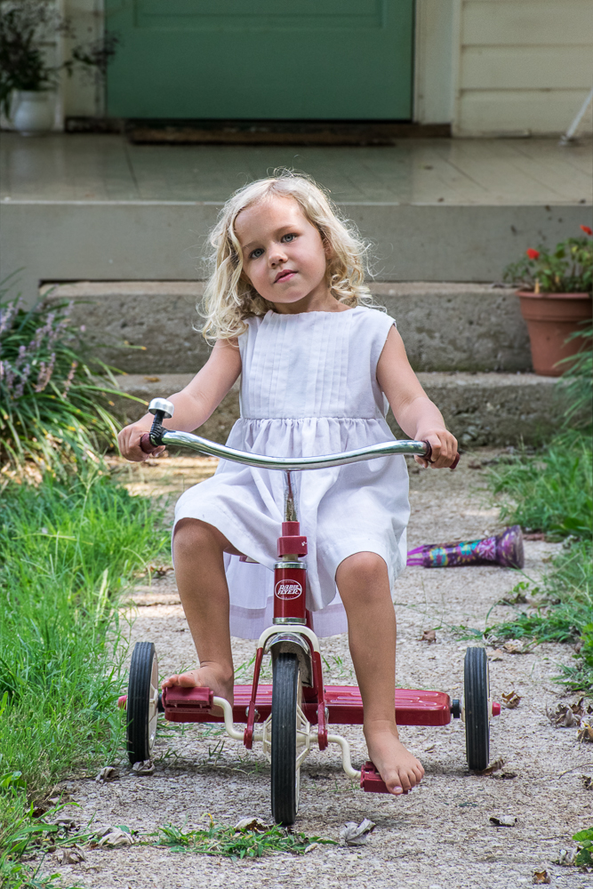 A little girl in a white dress riding a tricycle in front of her house.