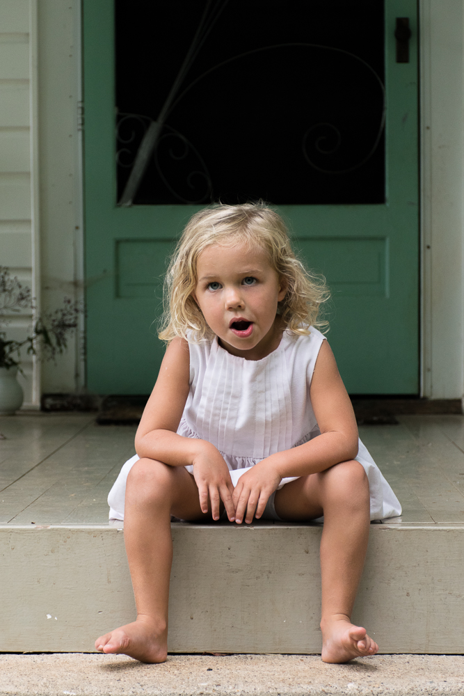 A little girl looking bored on the front porch of her farm house.