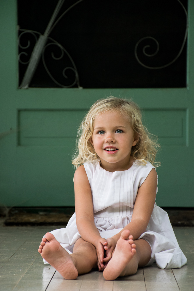 A little girl in a white dress sitting on the front porch of the farmhouse where she lives.
