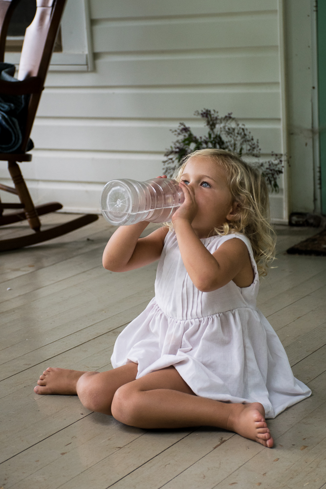 A little girl in a white dress sitting takes a big drink of water on her front porch.
