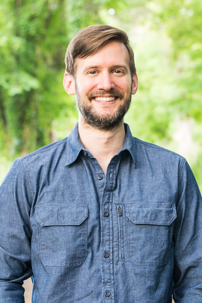 A man in a chambray shirt stands smiling in the woods.