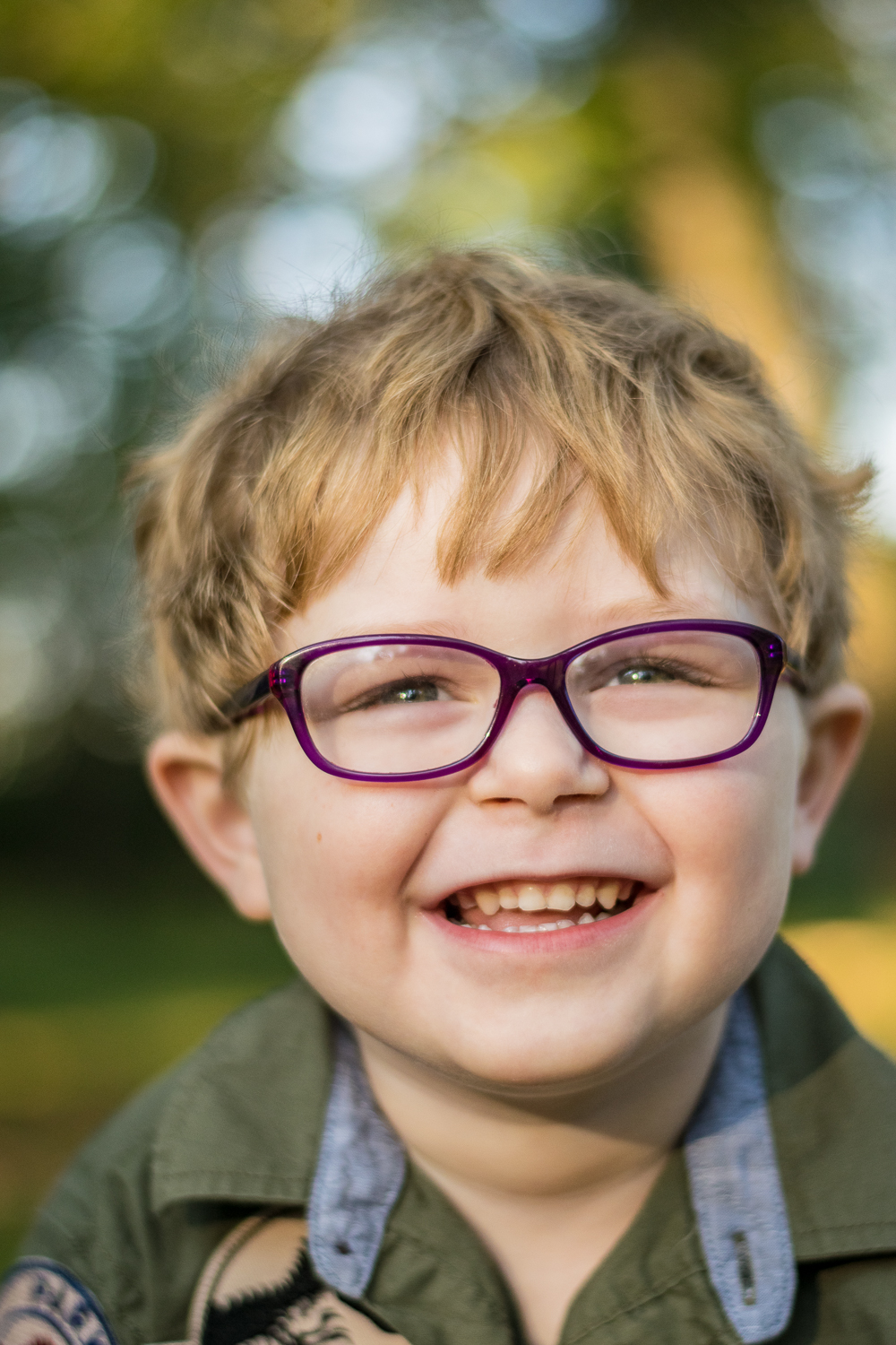 A close-up of a young boy with glasses laughing in the woods.
