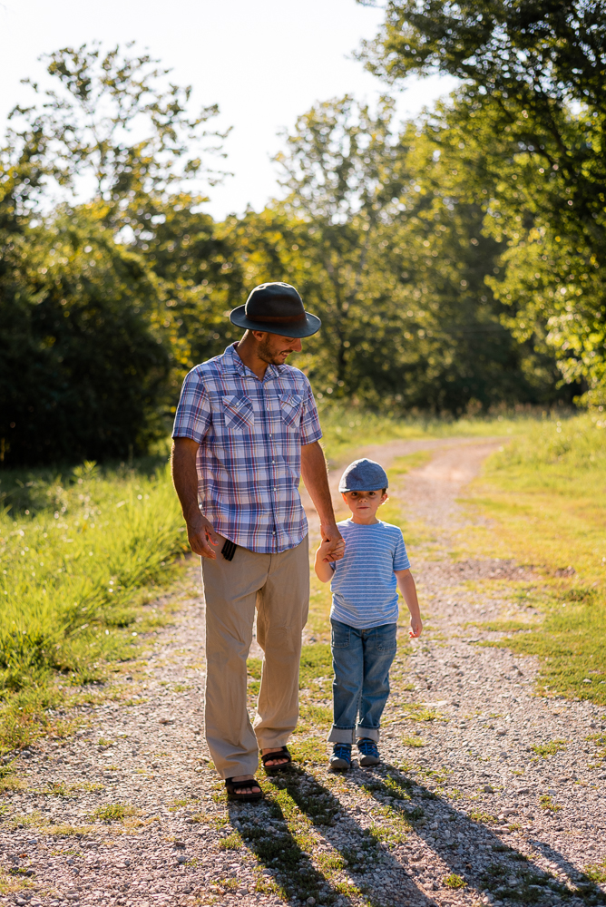 A father holds hands with his young son and walks down a gravel road.