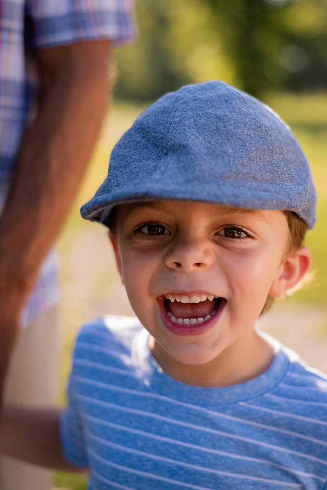 A little boy in a blue shirt and hat laughs at the camera.