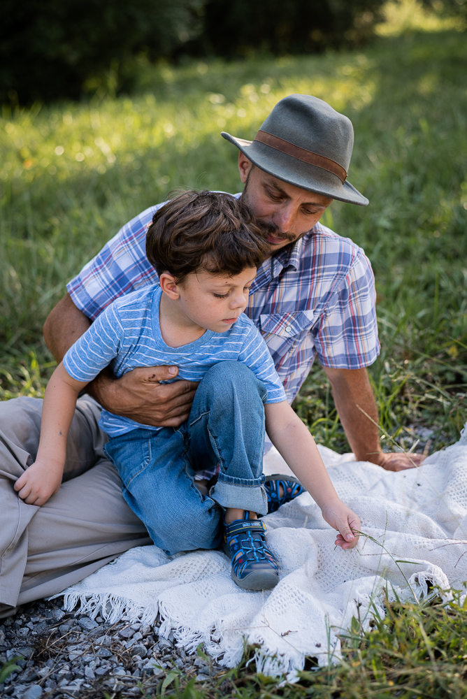 A father and son cuddle and sit on a blanket in the shade.