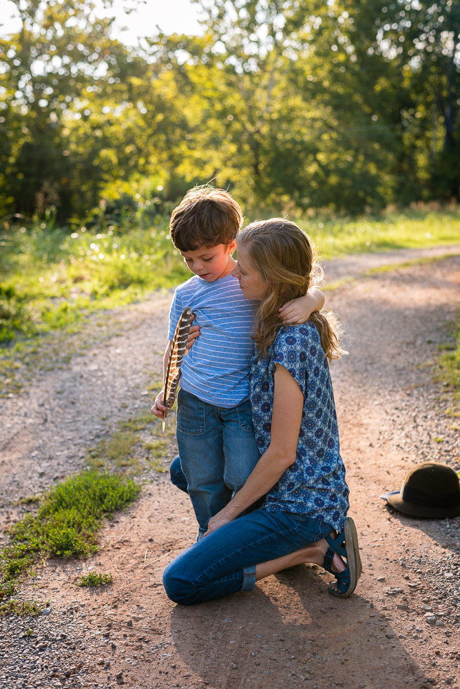 A kneeling mother and son hug in the sunshine on a gravel road.