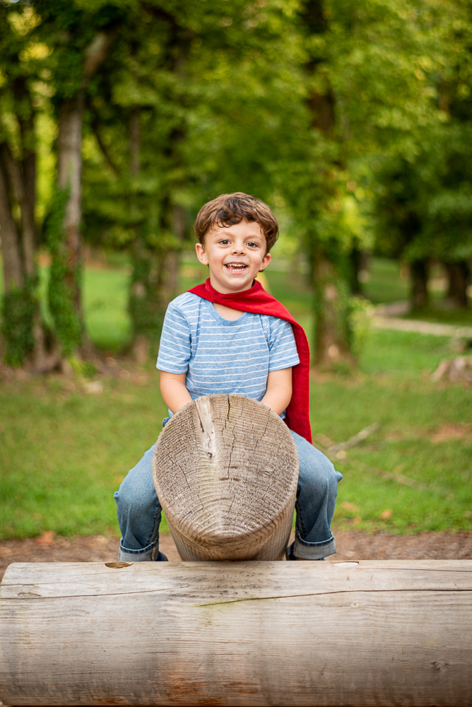A young boy wearing a red cape plays on a log play structure.