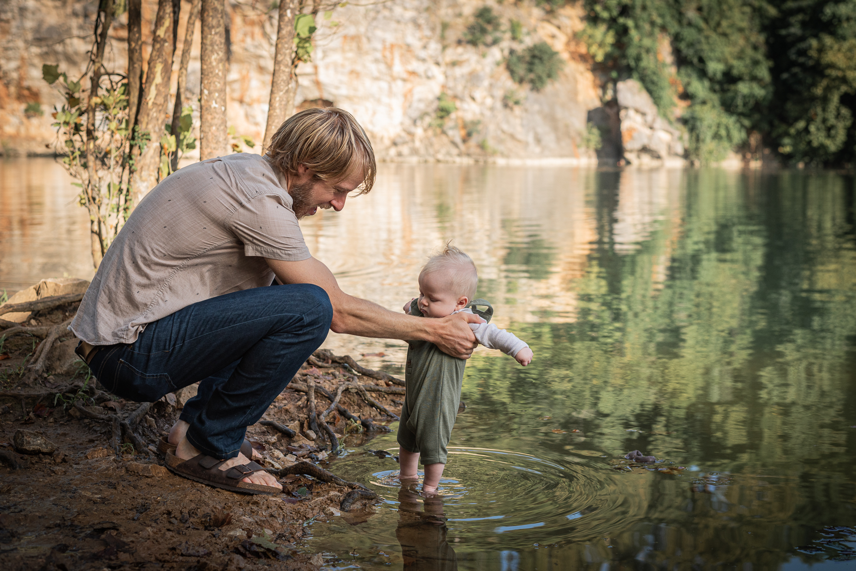 A father holds his baby boy's feet in the water at a quarry.