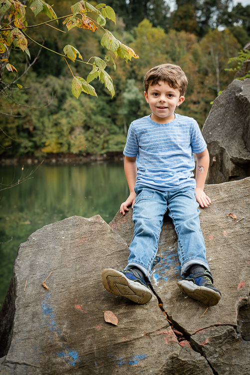 A little boy sits on top of a rock by a lake during the fall.