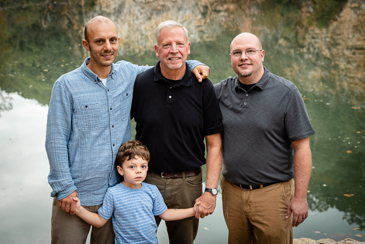 Three generations of men stand together in front of a lake.