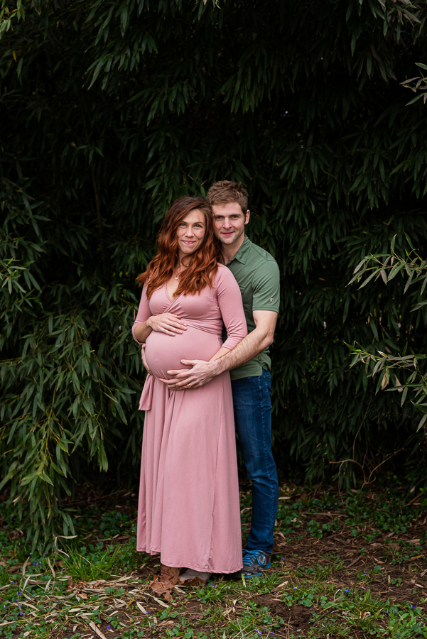 A pregnant woman and her husband embrace in a lush, green garden.