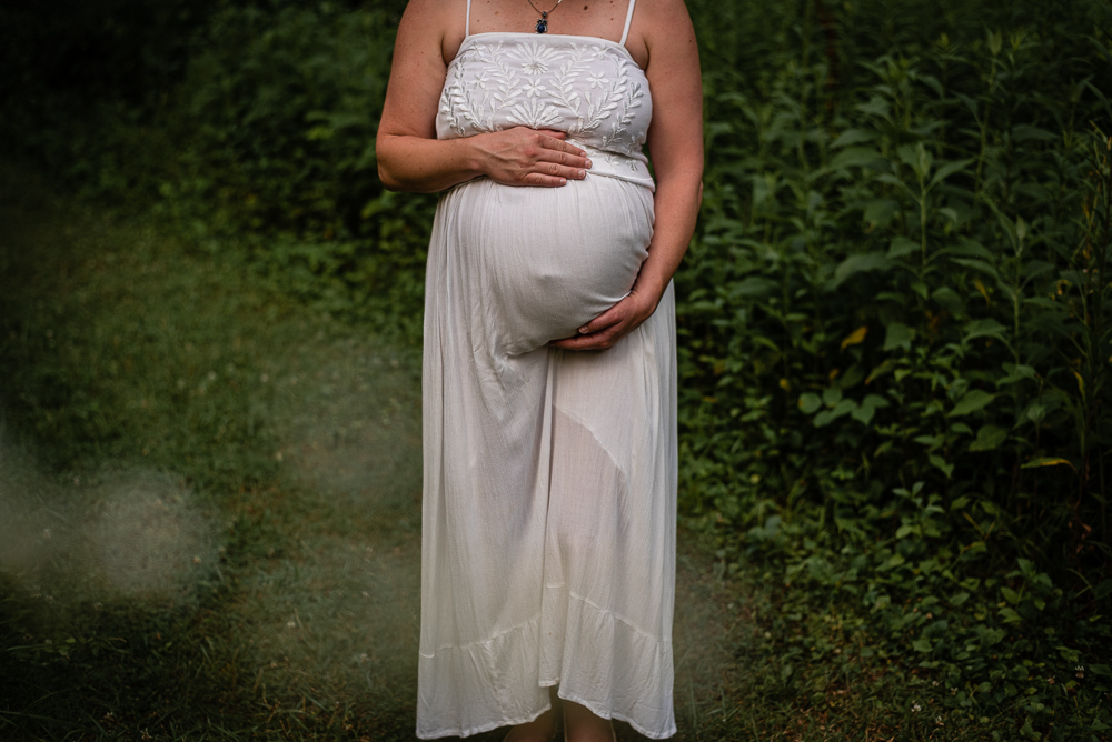 A cropped image of a pregnant woman in a white dress in a green field.