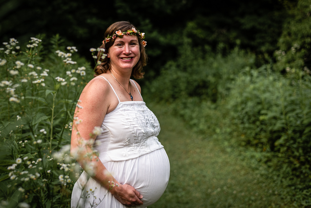 A pregnant woman in a white dress smiles and stands in a lush green field with flowers.