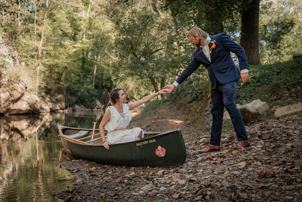 A groom reaches for his bride's hand to help her out of a canoe.