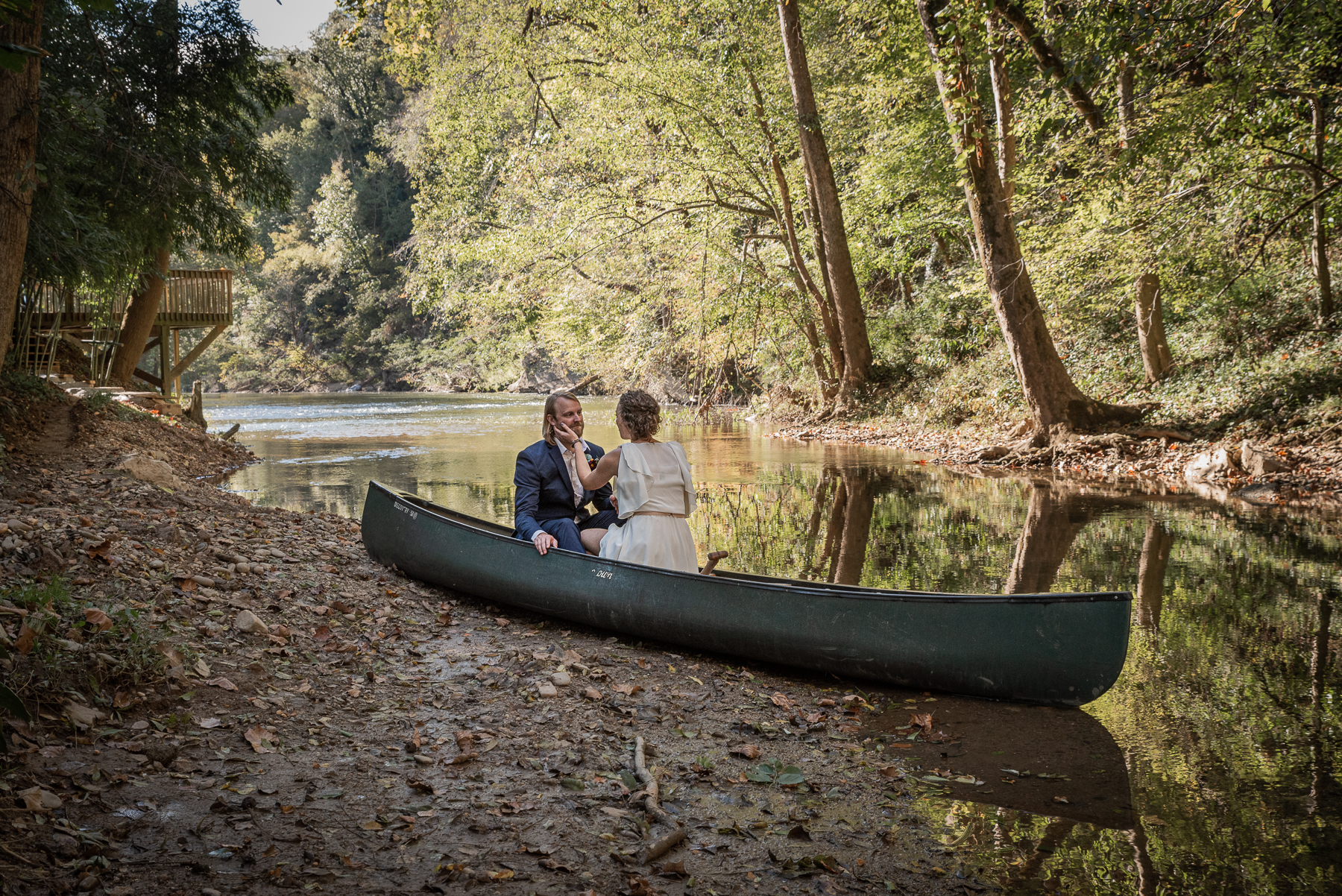 A bride and groom sit in a canoe on the bank of a creek.