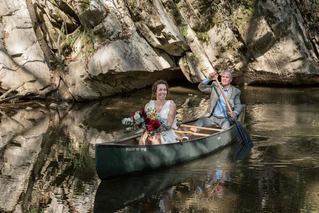 A bride and her father paddle down a creek in a canoe.