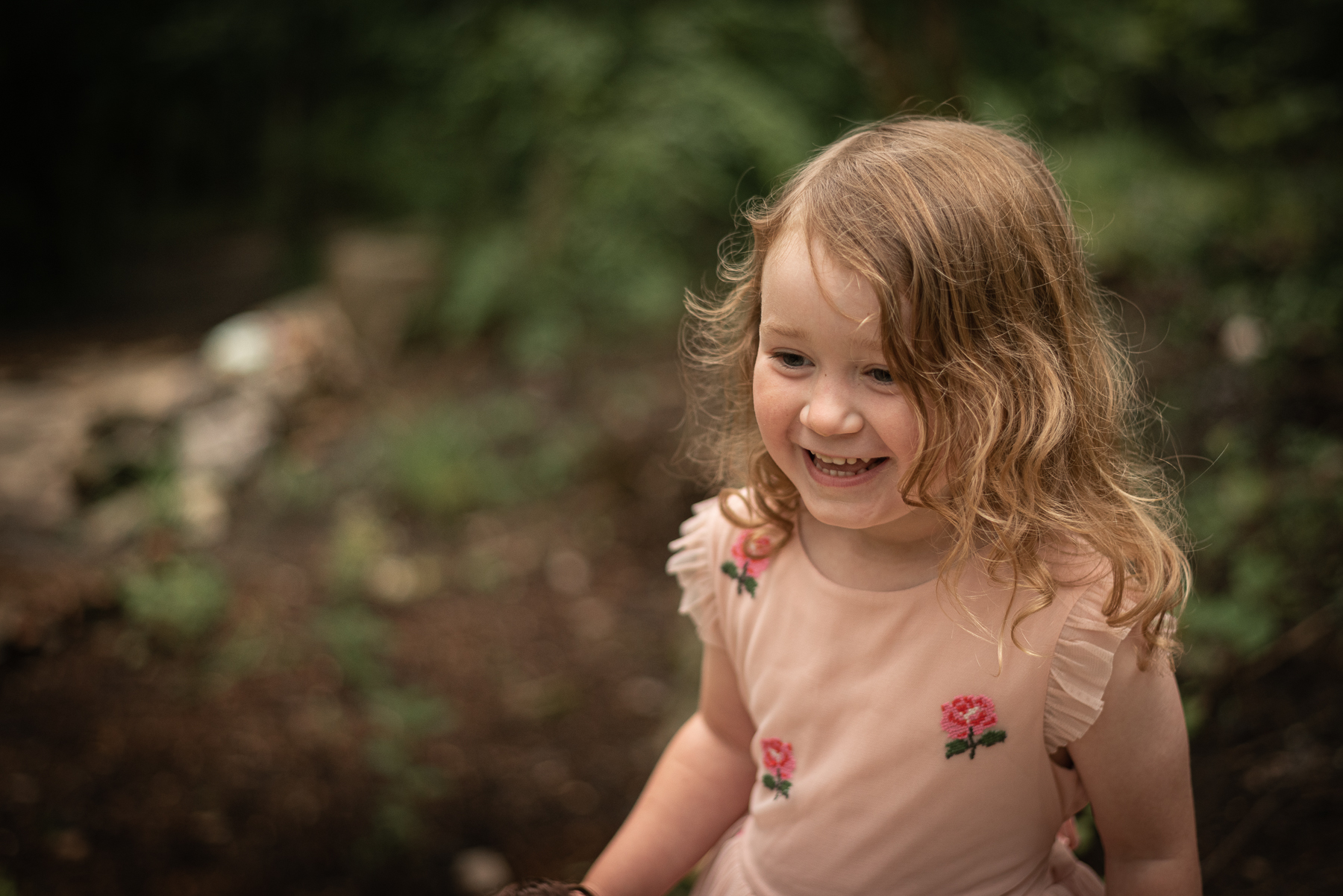 A portrait of a little girl in a pink dress smiling in the woods.