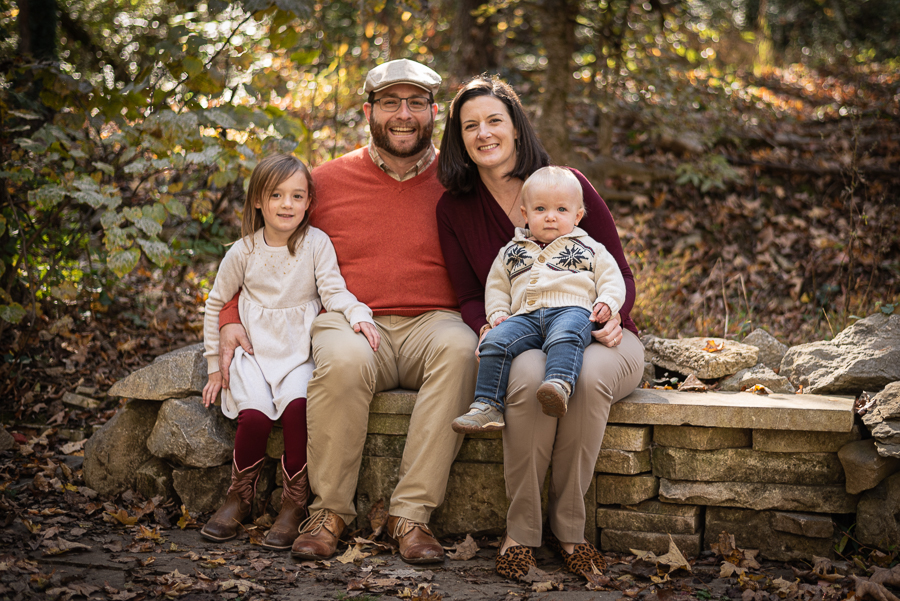 Parents and two young children sit on a stone wall in the forest.