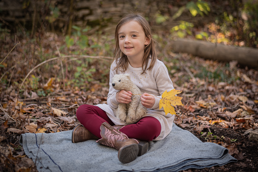 A young girl sits on the forest floor with her stuffed bear.