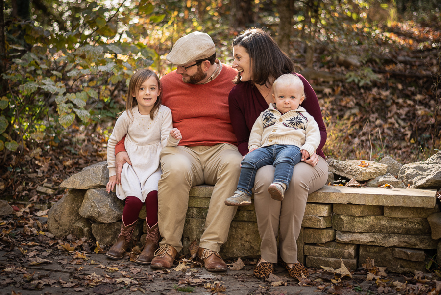 A young family of four sits on a stone wall in the forest.