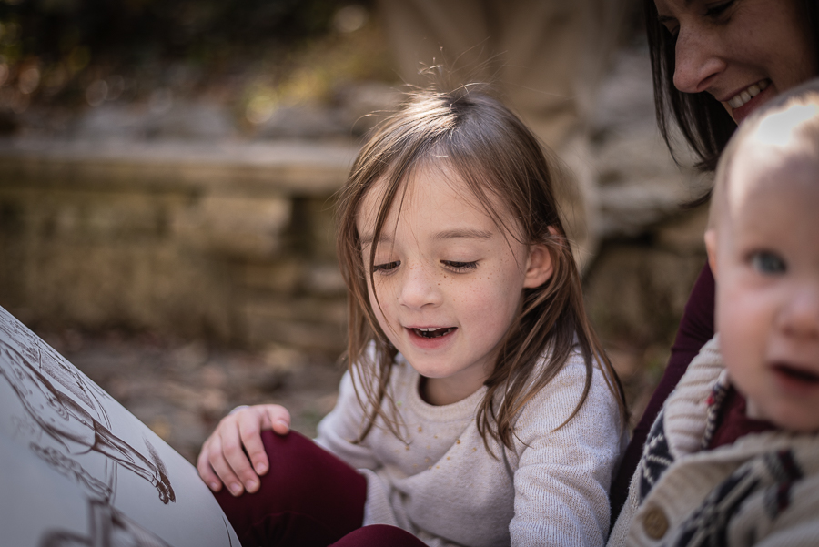 A young girl looks at a book in the woods with her family.